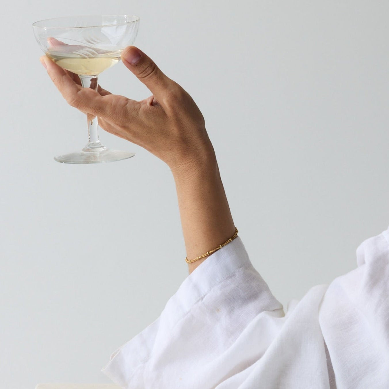 A person in a white long-sleeve top holds a clear coupe glass containing a pale yellow drink against a light gray background, with the word INDIA printed at the top.