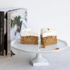 A styled kitchen scene with a white pedestal cake stand holding two slices of frosted cake, next to a stack of cookbooks and a dark brown ceramic mug filled with wooden spoons, all against a clean white wall and light marble surface.
