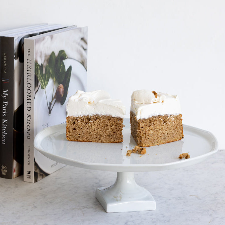A styled kitchen scene with a white pedestal cake stand holding two slices of frosted cake, next to a stack of cookbooks and a dark brown ceramic mug filled with wooden spoons, all against a clean white wall and light marble surface.