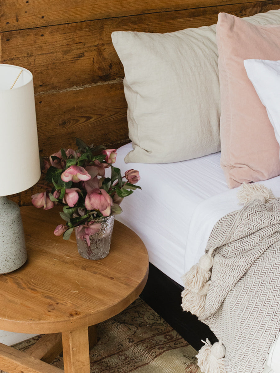 Bed with pillows and a blanket next to a wooden side table with a lamp and flowers.