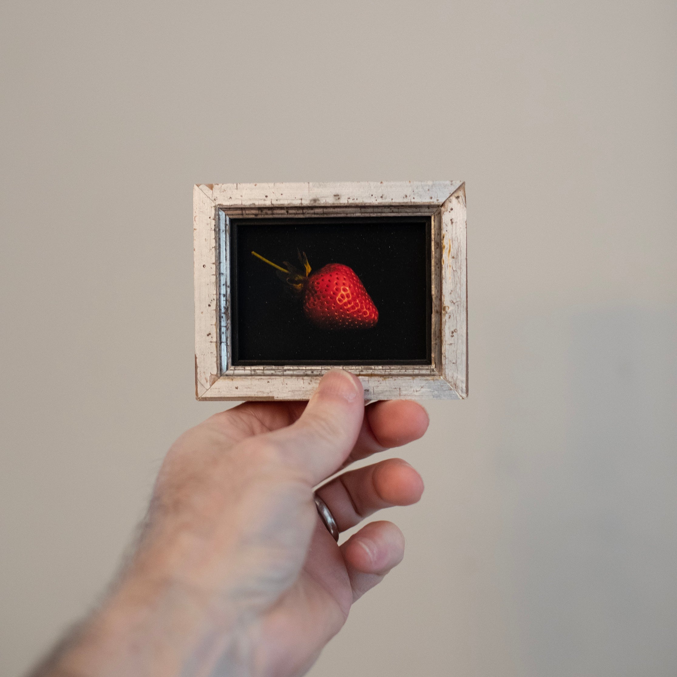Hand holding a small framed picture of a strawberry against a plain background